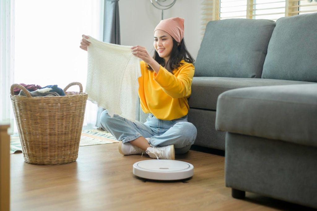 young-happy-woman-folding-clothes-living-room-while-robotic-vacuum-cleaner-working