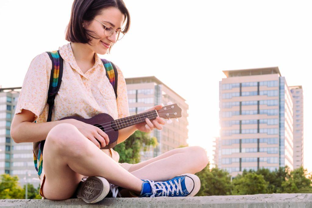 young smiling woman in sneakers playing ukulele at sunset in the city, amateur music concept and artistic lifestyle, copy space for text
