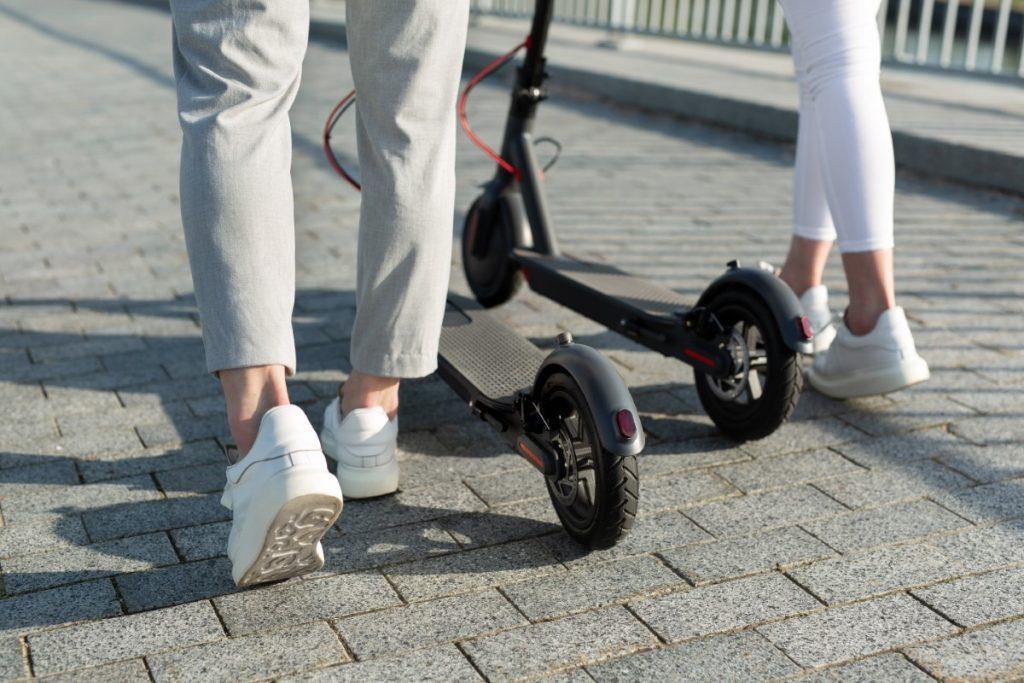 A couple of men and women are walking in the Park and pushing electric scooters